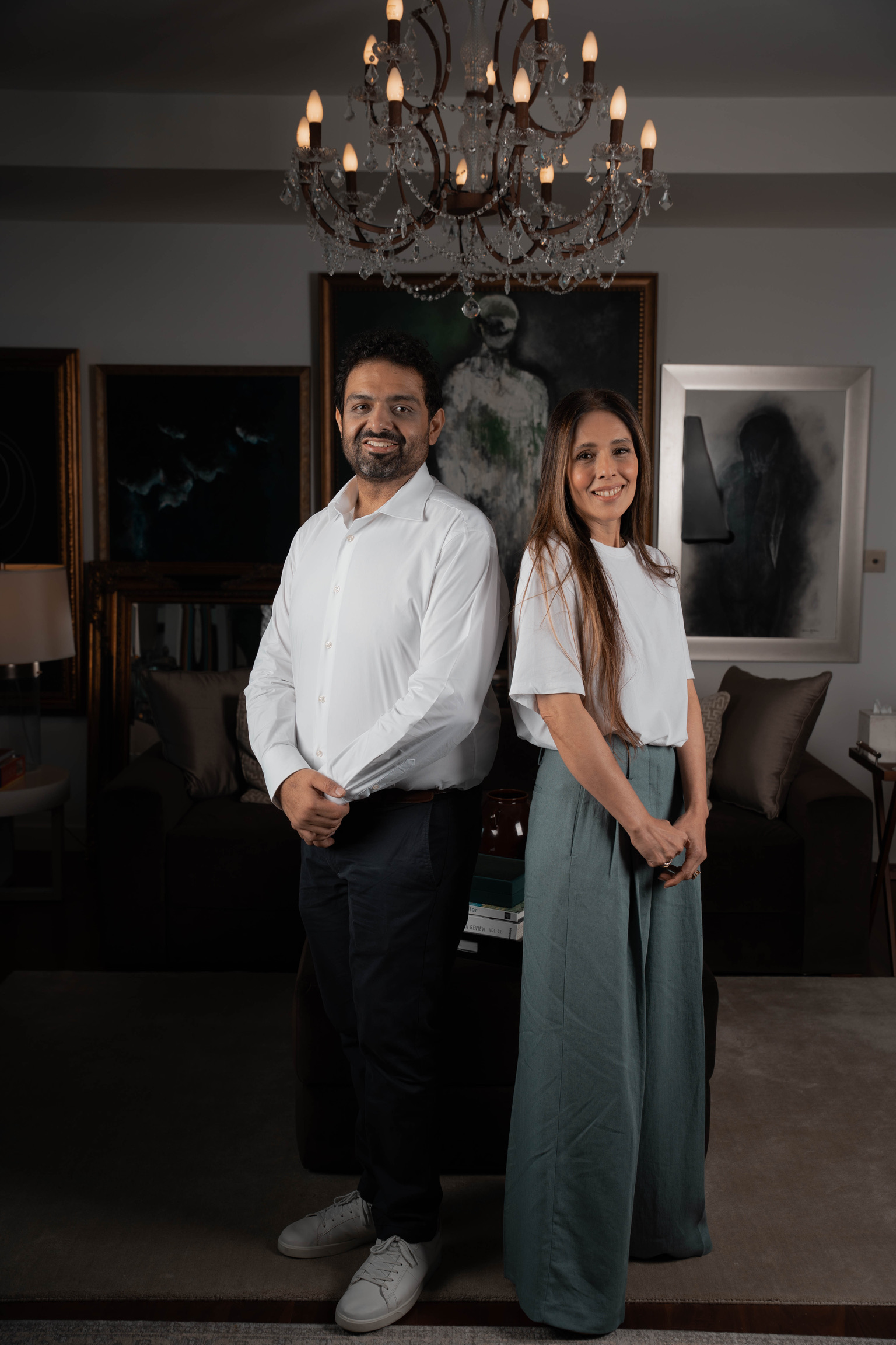 Bushra Khokhar and Tariq Ismail, co-founders of True Build Design, standing back to back beneath a chandelier in a collected interior