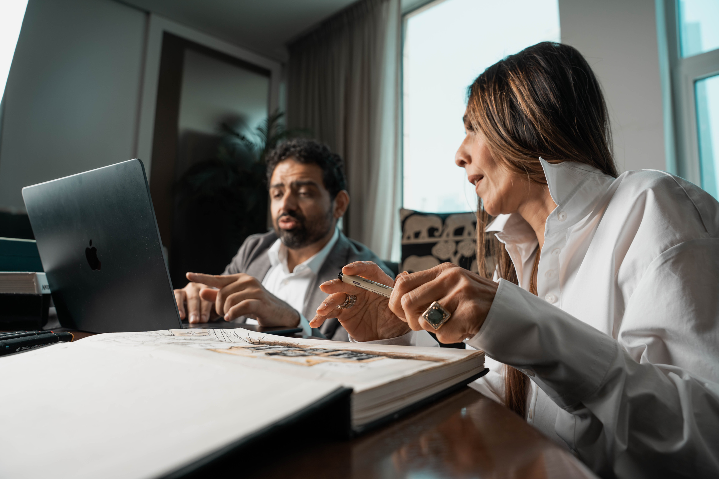 The founders working at a desk — her sketchbook and material sample, his laptop, the Dubai skyline in the window