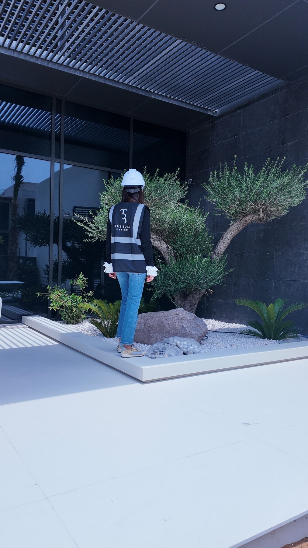 Bushra Khokhar in a True Build vest and hard hat on site, studying an olive tree in a courtyard against a dark stone wall